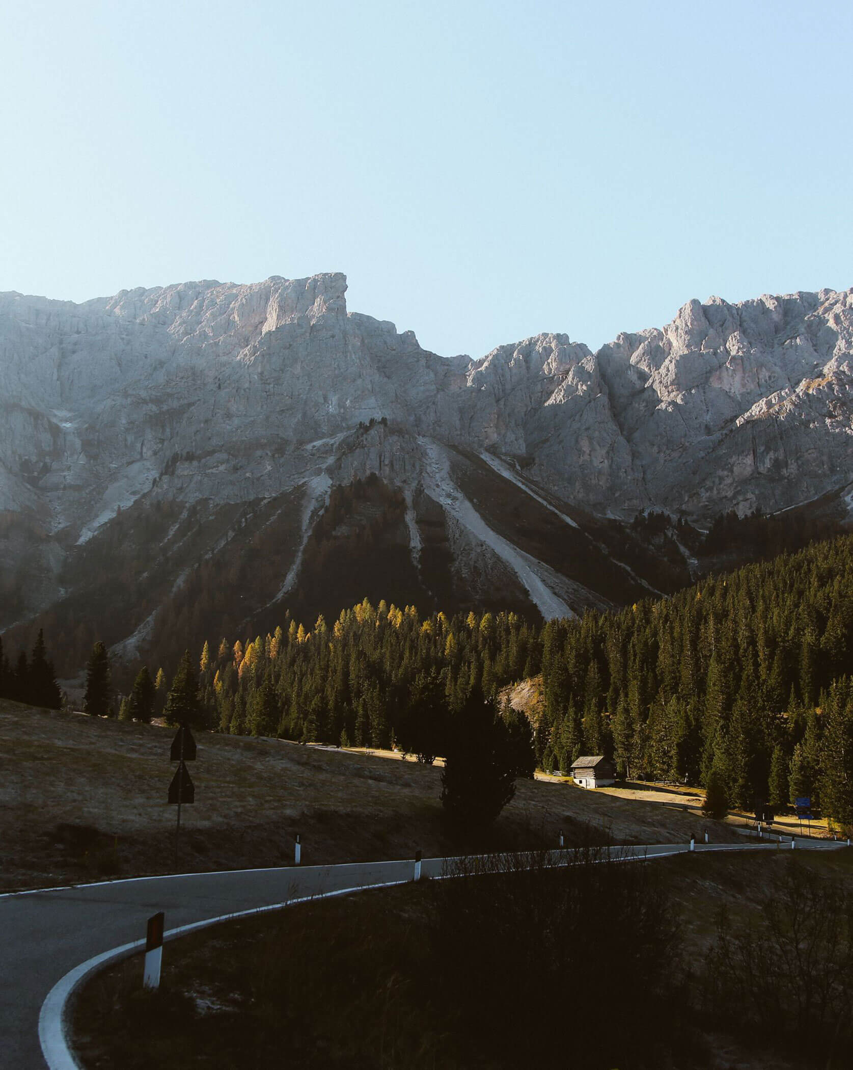 Eine kurvenreiche Straße vor Kiefern und hohen, felsigen Bergen unter einem klaren Himmel. - Lüch de Poldele