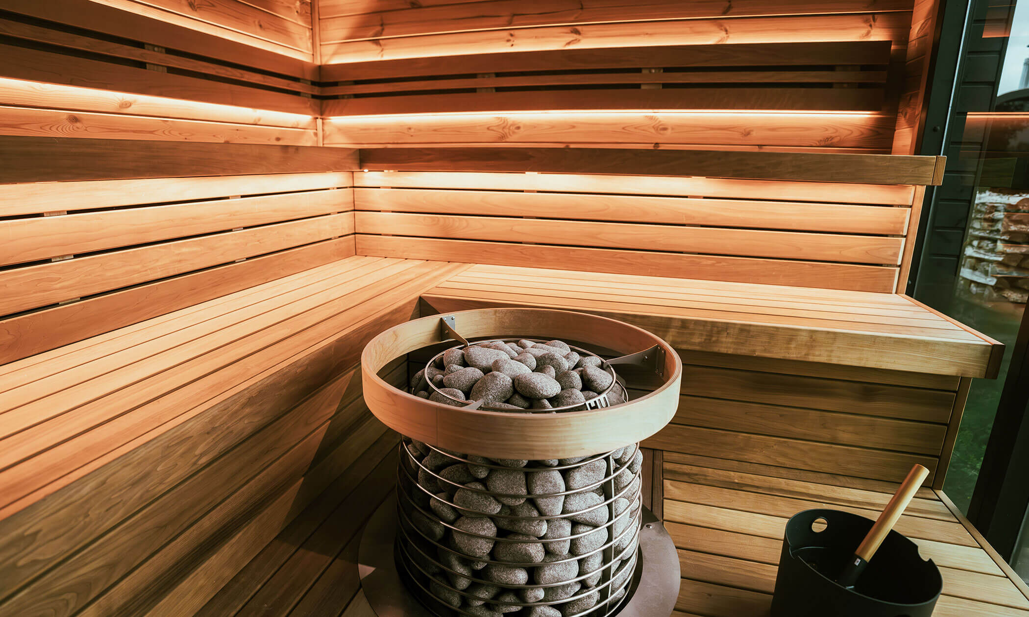 Innenraum einer Holzsauna mit heißen Steinen im Ofen und einem schwarzen Eimer mit einer Schöpfkelle auf der Bank. - Lüch de Poldele