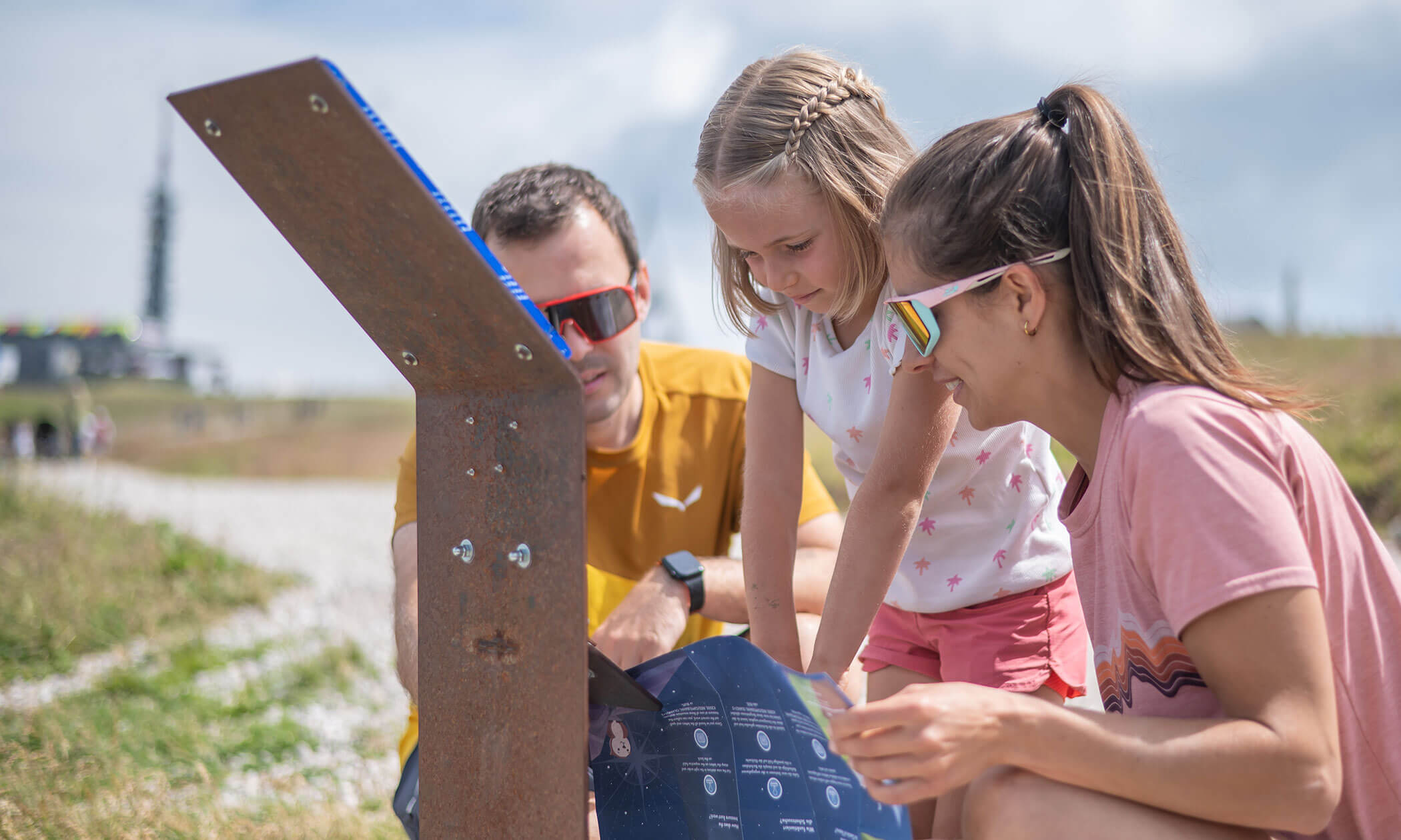 Drei Personen, die im Freien eine Karte und eine Informationstafel betrachten; alle tragen an einem sonnigen Tag eine Sonnenbrille. - Lüch de Poldele