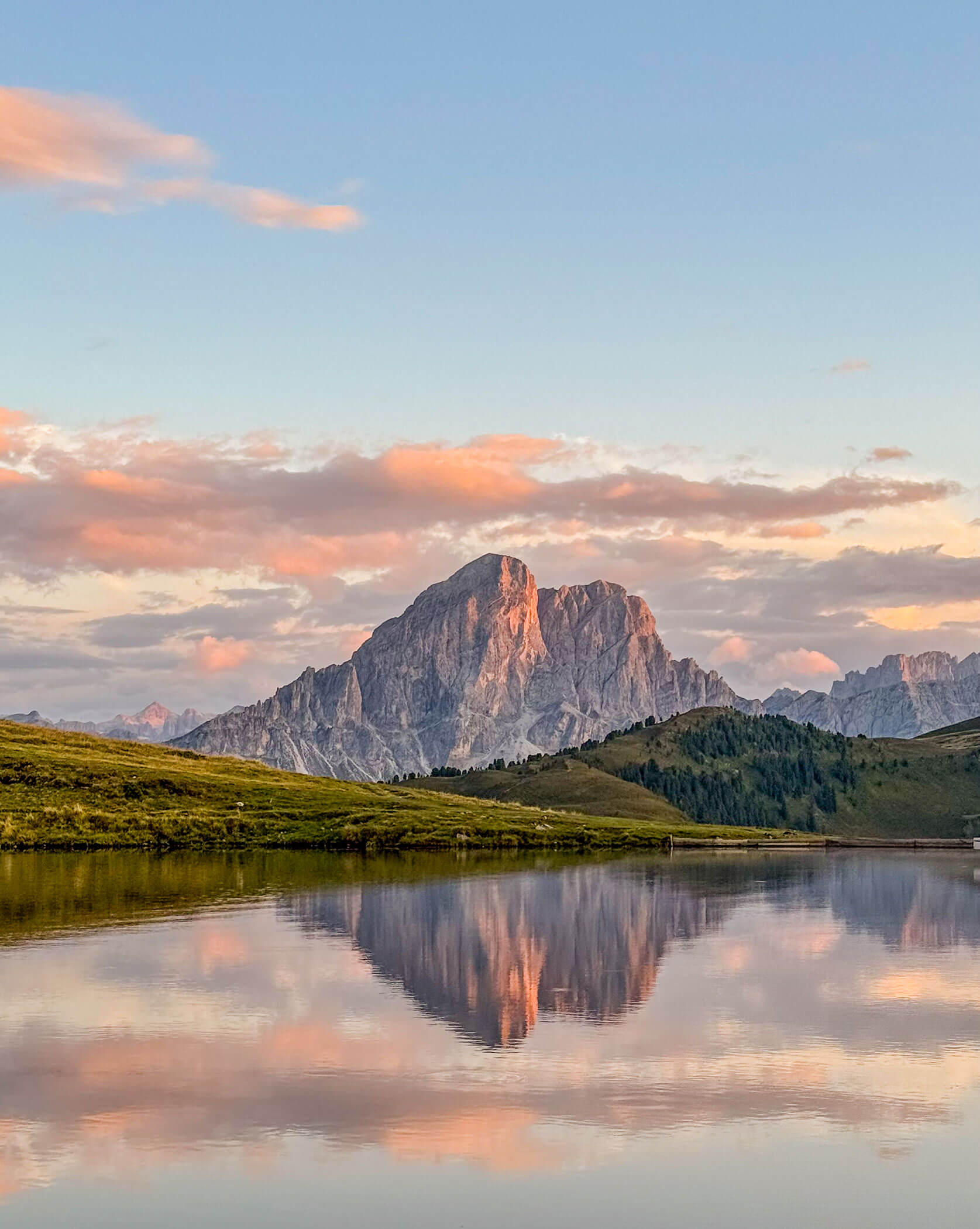 Ein hoher Berg spiegelt sich bei Sonnenuntergang in einem ruhigen See wider, während am Himmel rosa Wolken zu sehen sind. - Lüch de Poldele