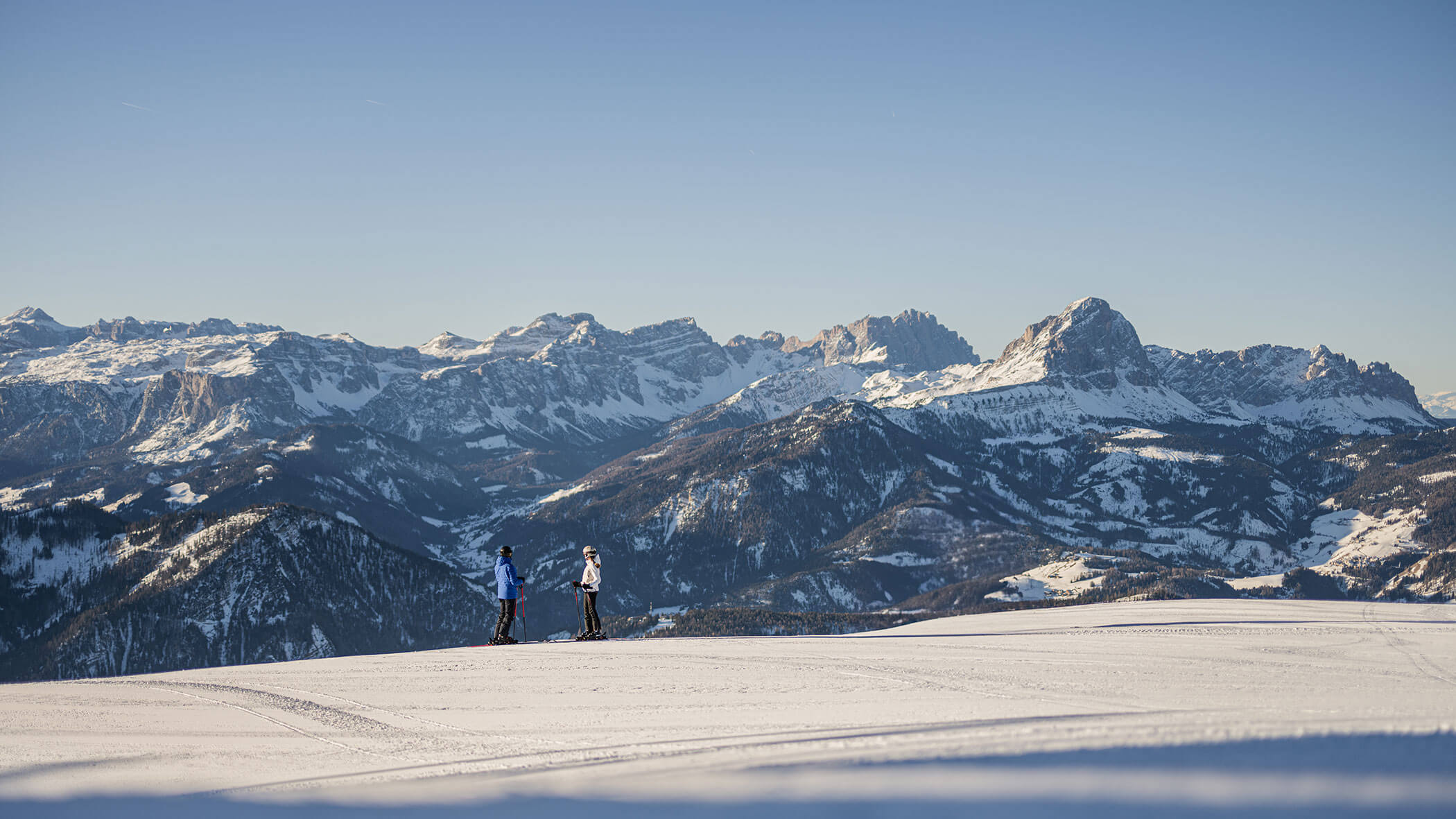 Zwei Menschen, die auf einem verschneiten Hang Ski fahren, im Hintergrund sind Berge zu sehen, über ihnen erstreckt sich ein strahlend blauer Himmel. - Lüch de Poldele