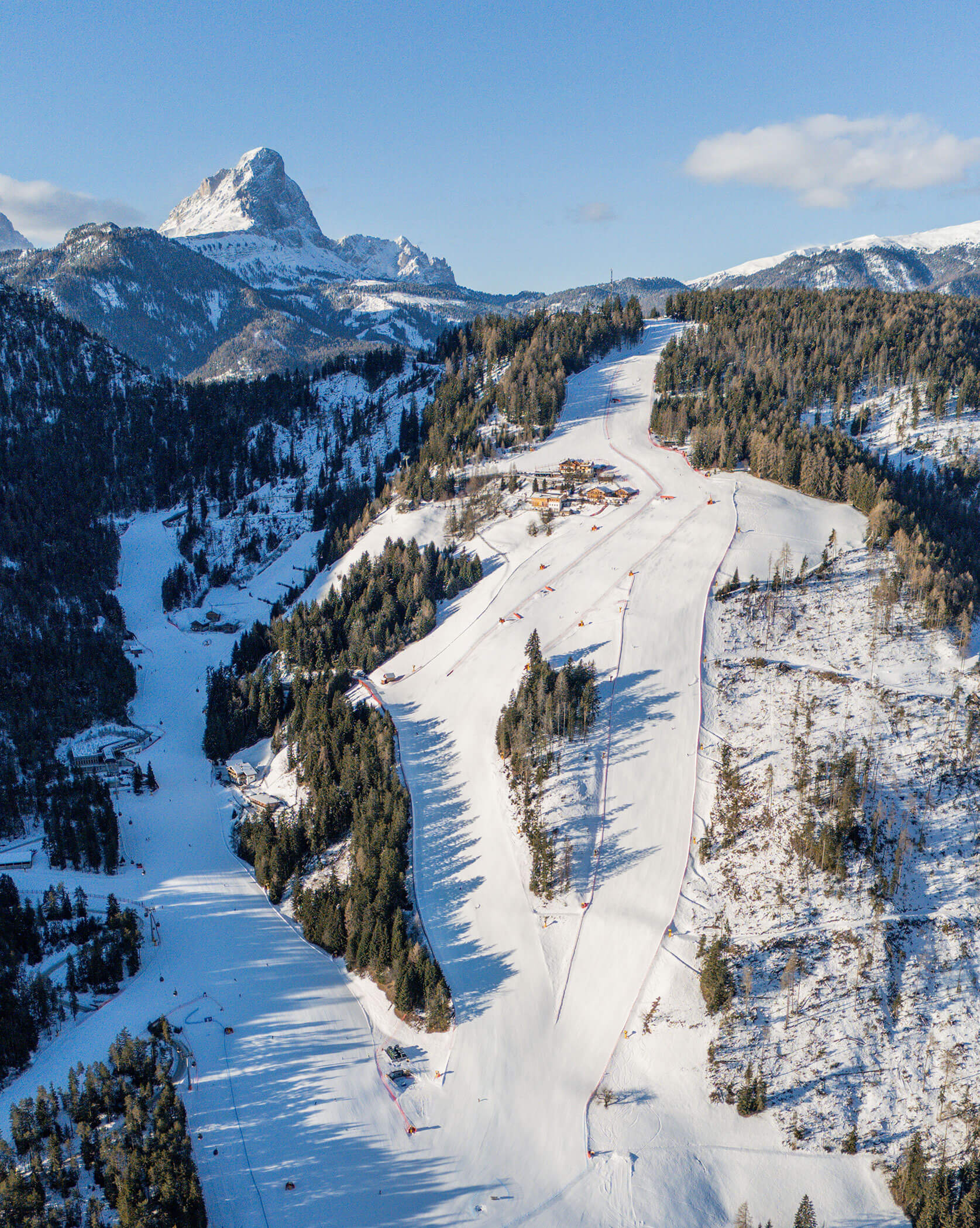 Luftaufnahme einer verschneiten Skipiste, umgeben von Bäumen, mit Bergen im Hintergrund unter blauem Himmel. - Lüch de Poldele