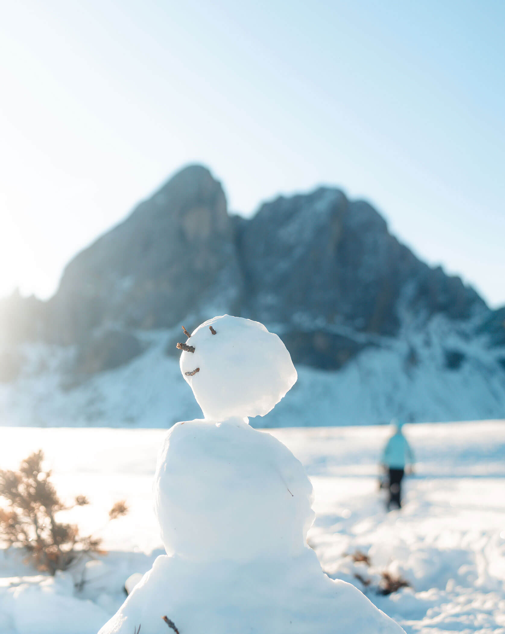 Ein kleiner Schneemann mit Stöckchenarmen steht auf einem verschneiten Feld, im Hintergrund sind Berge und eine Person zu sehen. - Lüch de Poldele