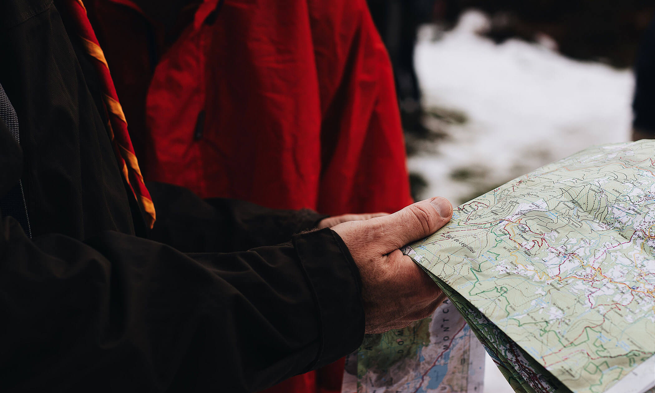 Eine Person in einer dunklen Jacke, die im Freien eine Karte in der Hand hält, daneben eine weitere Person in einer roten Jacke. Auf dem Boden liegt Schnee. - Lüch de Poldele