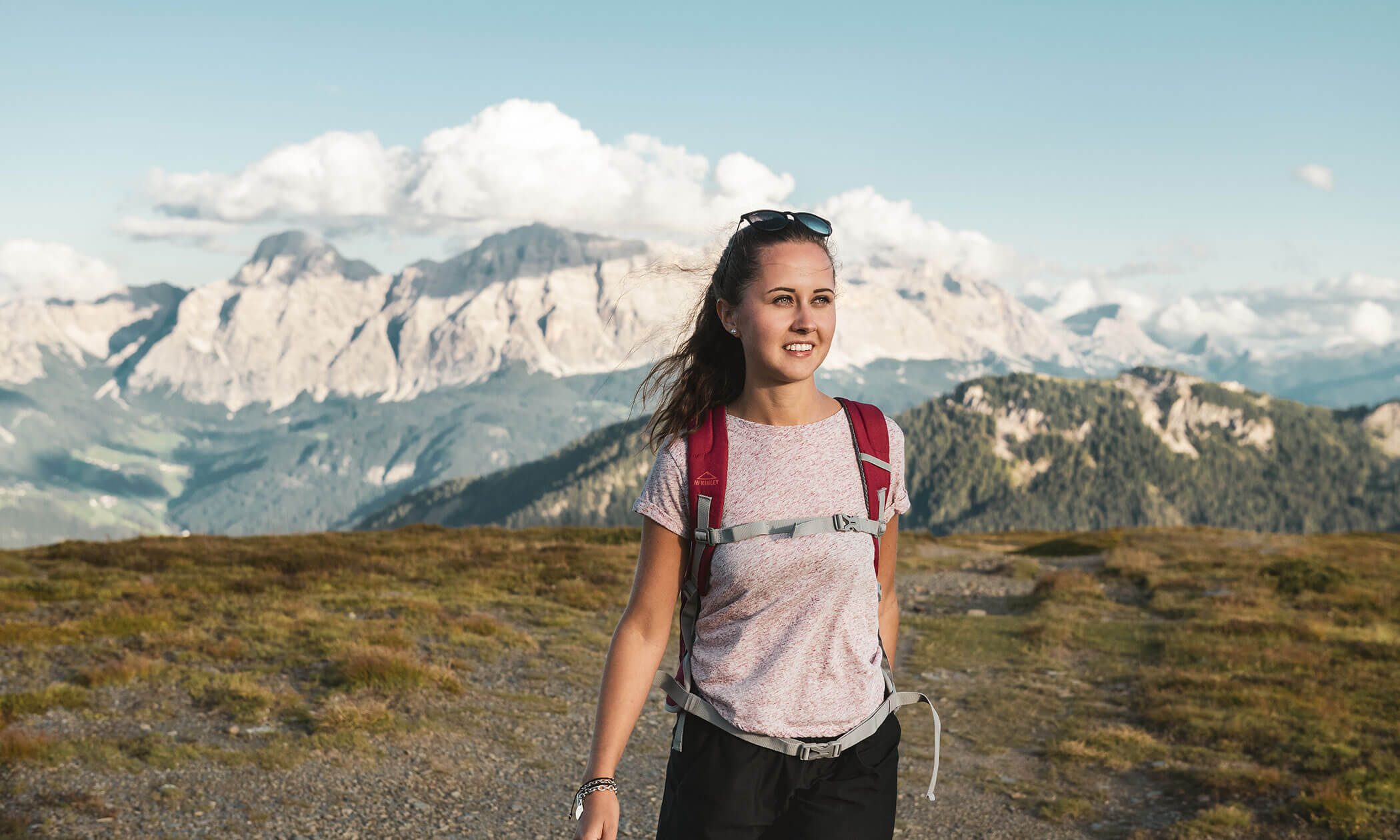 Eine Frau mit Rucksack wandert auf einem grasbewachsenen Berg, im Hintergrund sind entfernte Gipfel und ein blauer Himmel zu sehen. - Lüch de Poldele
