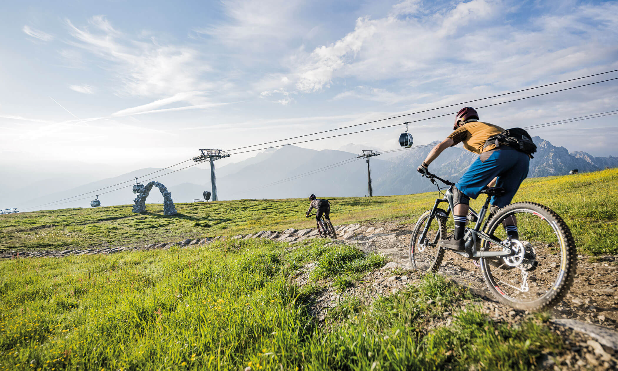Zwei Radfahrer fahren mit ihren Mountainbikes auf einem grasbewachsenen Weg in der Nähe von Seilbahnen und Bergen unter blauem Himmel. - Lüch de Poldele