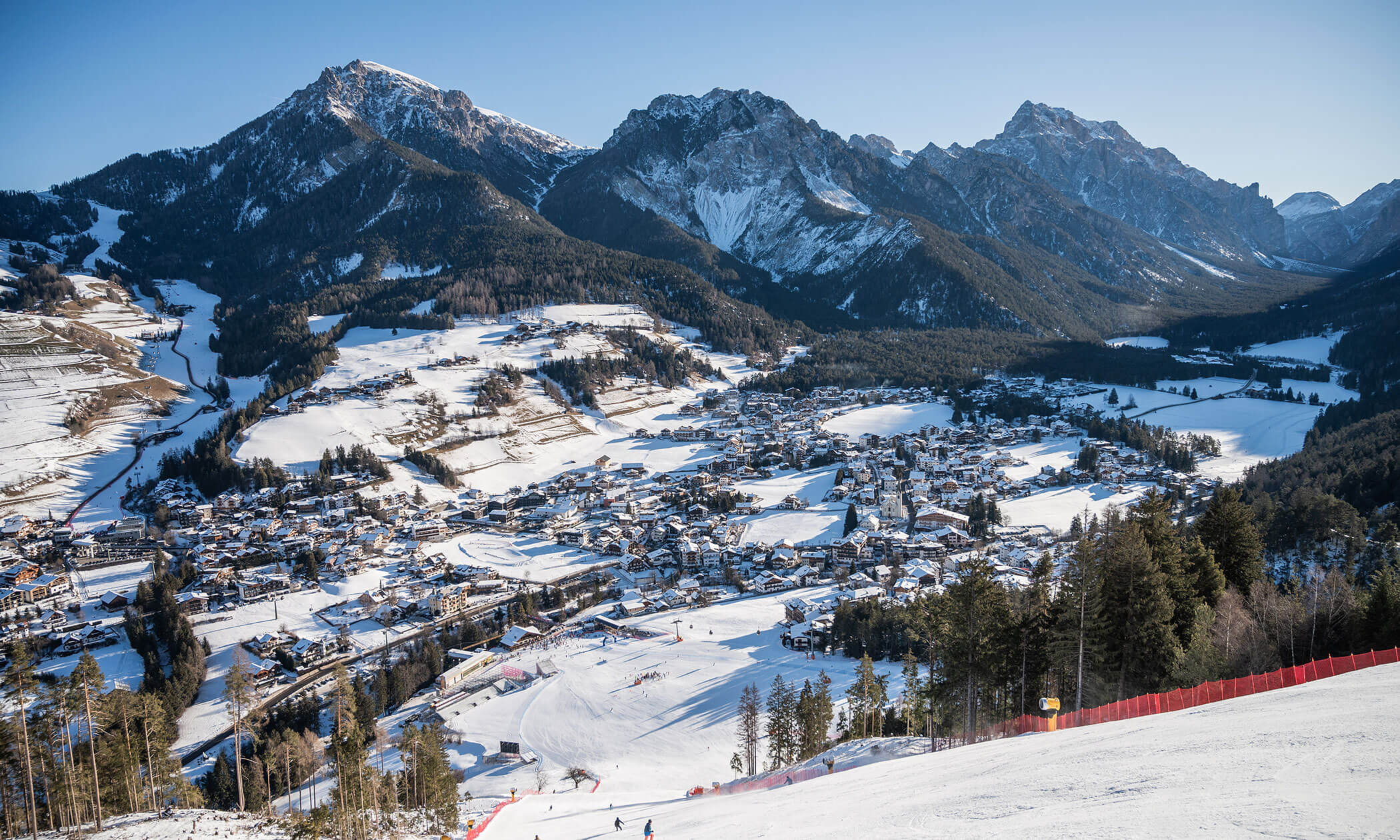 Ein schneebedecktes Dorf in einem Tal, umgeben von hohen Bergen unter einem strahlend blauen Himmel, von einer Skipiste aus gesehen. - Lüch de Poldele
