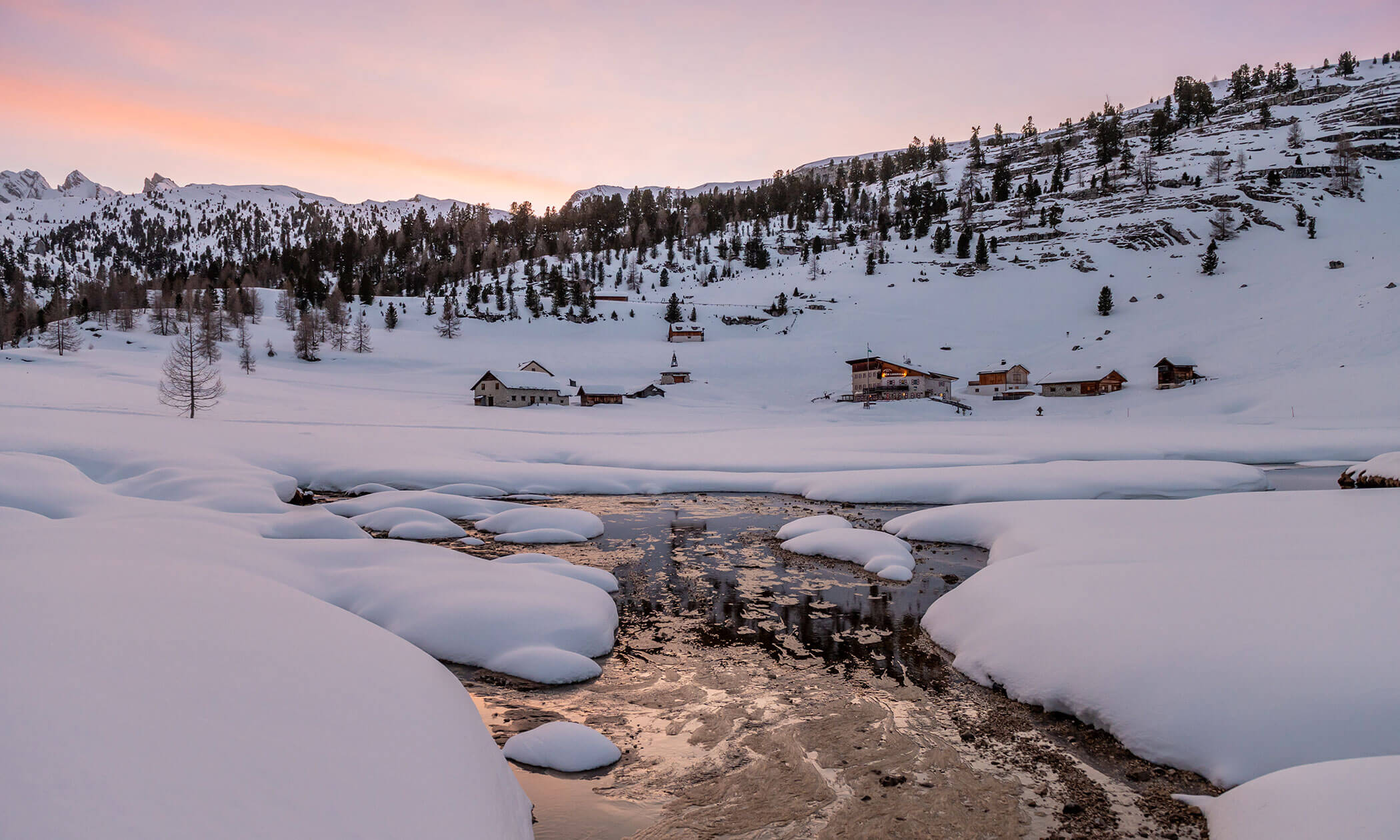 Ein verschneites Dorf bei Sonnenuntergang mit einem Bach, schneebedeckten Bäumen, Häusern und Bergen im Hintergrund. - Lüch de Poldele
