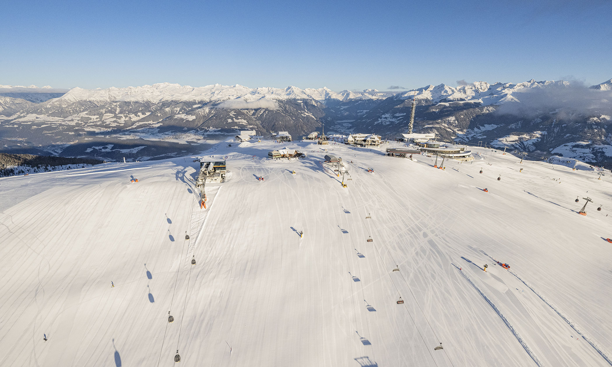 Luftaufnahme einer verschneiten Skipiste mit Skiliften, Gebäuden und Bergen in der Ferne unter klarem Himmel. - Lüch de Poldele